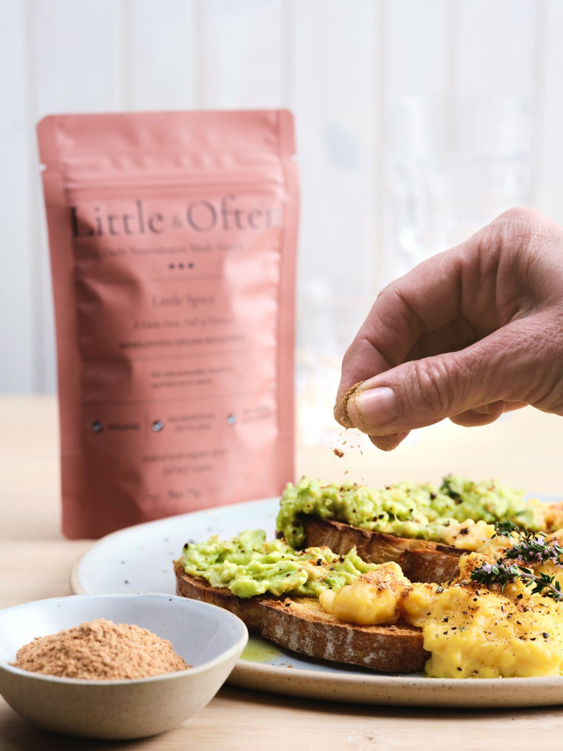 Hand sprinkling Little & Often Little Spice seasoning over avocado toast and scrambled eggs, with the pouch in the background.