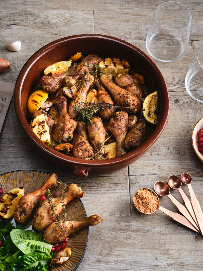Roasted chicken drumsticks with lemon and herbs in a brown ceramic dish, beside a plate of chicken and salad.